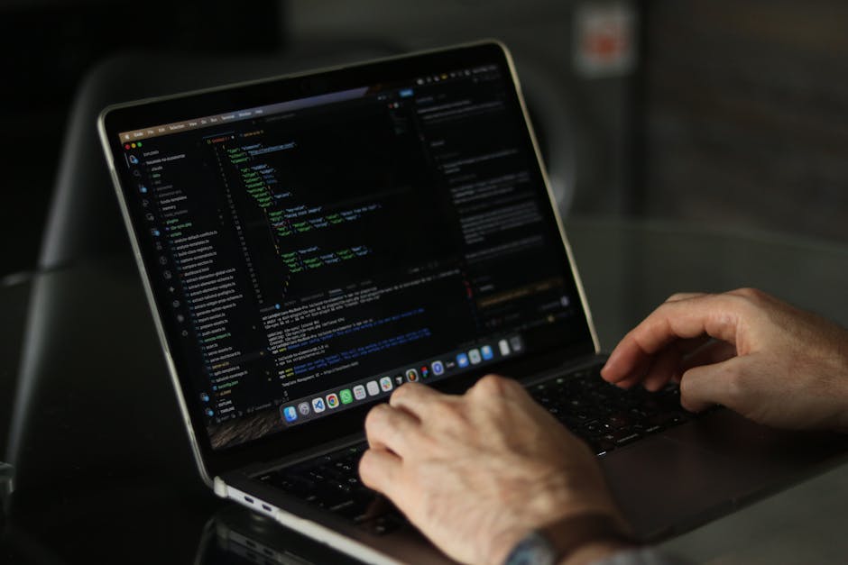 A person typing on a laptop with a black screen displaying lines of colorful code, situated on a dark, reflective surface. The scene is illuminated by soft ambient lighting, highlighting the keyboard 