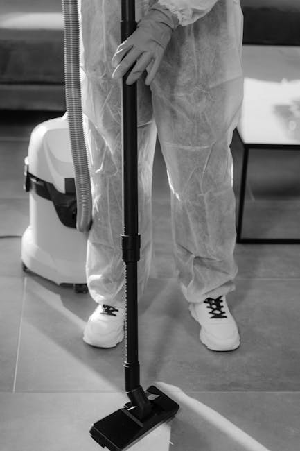 A person dressed in protective white coveralls, gloves, and sneakers is using a handheld vacuum cleaner with a long hose attachment to clean a hard floor surface. The scene likely takes place in a cle